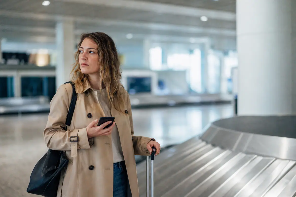 Voyageuse consultant son téléphone près d’un tapis à bagages vide dans un hall d’arrivée d’aéroport