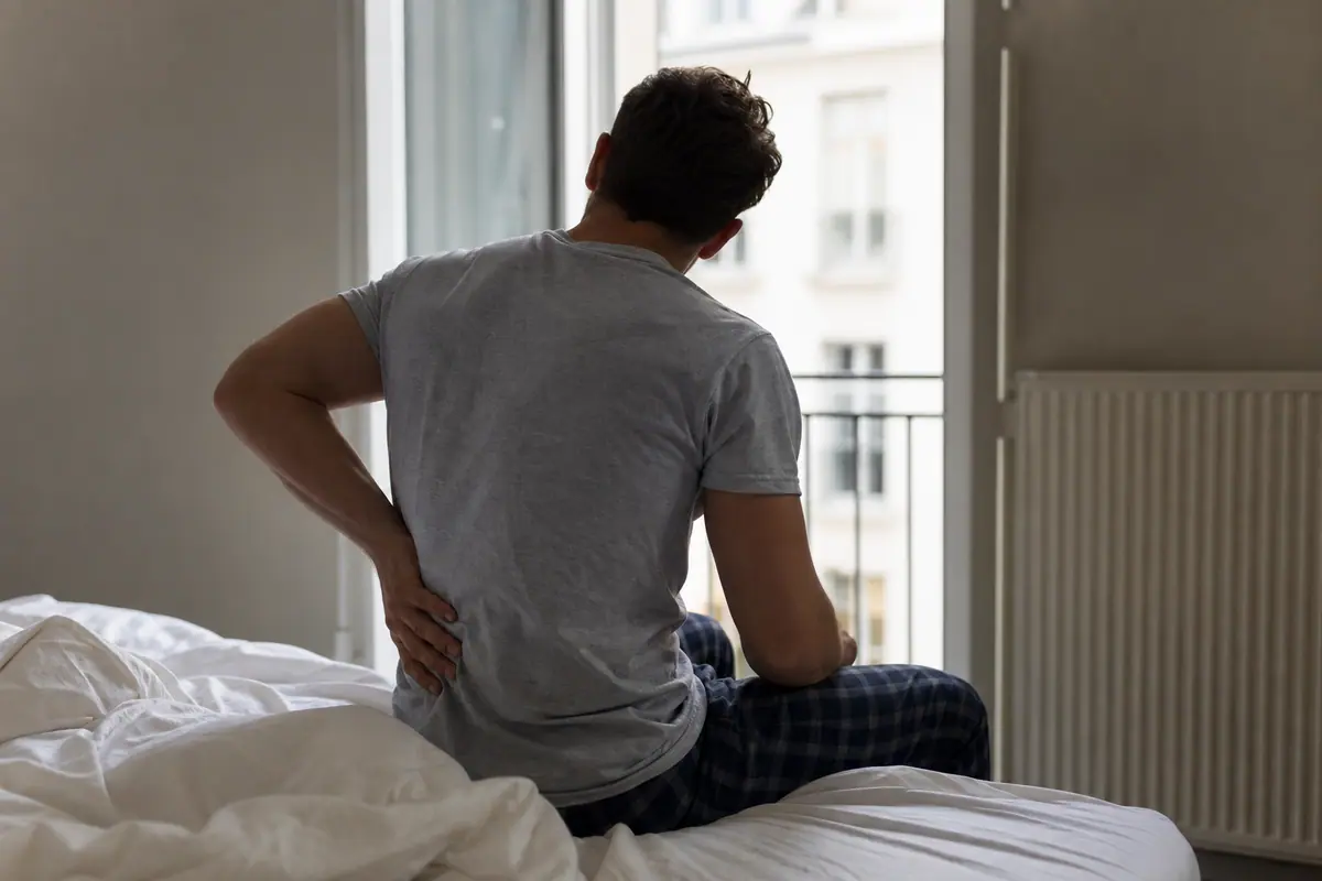 Personne qui s’étire le dos assise au bord du lit au réveil, lumière du matin